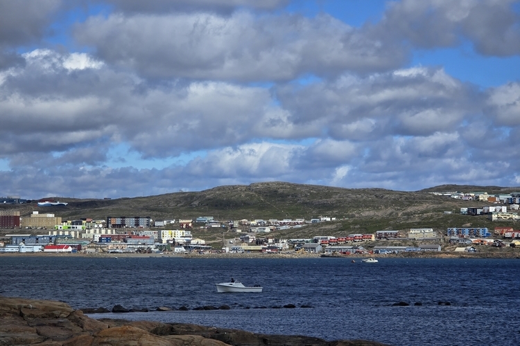 View of Iqaluit, Nunavut from across the bay in summer. The sky is blue with many clouds. There is a motorboat in the water.