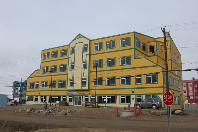 A four-storey yellow building with green window frames. A stop sign is visible in the lower right corner with writing in English and Inuktitut.