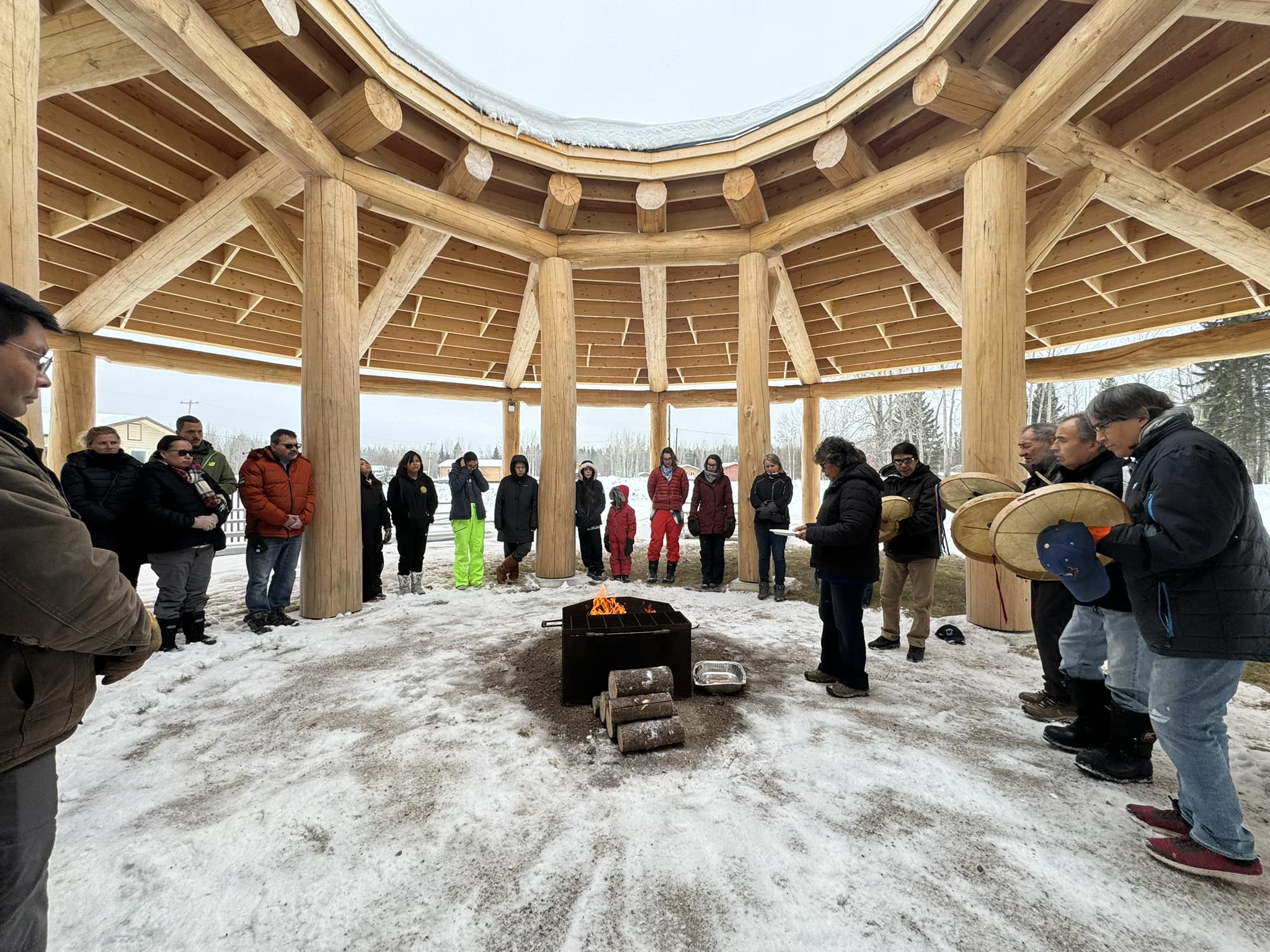 Des adultes et des enfants vêtus d’habits de neige se rassemblent autour d’un petit feu à l’intérieur d’une structure en bois composée de deux cercles de pôles externes et internes. Les poutres, qui s’étendent des poteaux extérieurs aux poteaux intérieurs, soutiennent un toit en angle avec une grande ouverture centrale. Les personnes se tiennent en cercle sous le toit et le feu brûle sous l’ouverture. La neige saupoudre le sol et quatre personnes jouent du tambour à main.